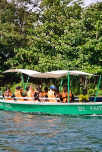 A group of people wearing orange life vests are seated in a green motorboat with a white canopy. The boat is on a body of water and is surrounded by lush green trees. Some of them appear to be enjoying the ride, and there is text on the boat's hull that reads 'BY FAITH.'