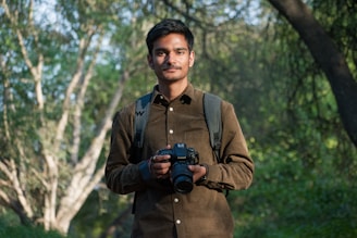 A person stands outdoors holding a camera, dressed in a brown shirt and a backpack, surrounded by greenery and trees in a natural setting.