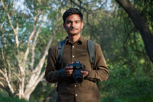 A person stands outdoors holding a camera, dressed in a brown shirt and a backpack, surrounded by greenery and trees in a natural setting.
