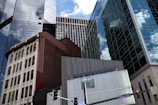 Tall, modern skyscrapers with reflective glass windows stand against a blue sky with scattered clouds. In the foreground, a classic older building with ornate architecture contrasts sharply with the contemporary structures. A traffic light and street signs marked Liberty Ave and Market St can be seen at the bottom of the image.