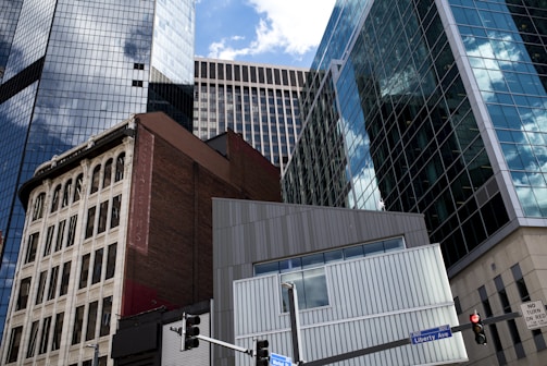 Tall, modern skyscrapers with reflective glass windows stand against a blue sky with scattered clouds. In the foreground, a classic older building with ornate architecture contrasts sharply with the contemporary structures. A traffic light and street signs marked Liberty Ave and Market St can be seen at the bottom of the image.