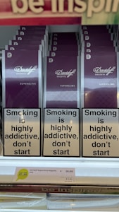 Several packs of Davidoff Magenta Superslims cigarettes are displayed on a shelf. Prominently, there are warning labels stating 'Smoking is highly addictive, don&rsquo;t start.' The price tag below shows &euro;39.00 for a carton and &euro;3.90 per pack. At the top, part of another sign is partially visible.