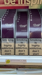 Several packs of Davidoff Magenta Superslims cigarettes are displayed on a shelf. Prominently, there are warning labels stating 'Smoking is highly addictive, don’t start.' The price tag below shows €39.00 for a carton and €3.90 per pack. At the top, part of another sign is partially visible.