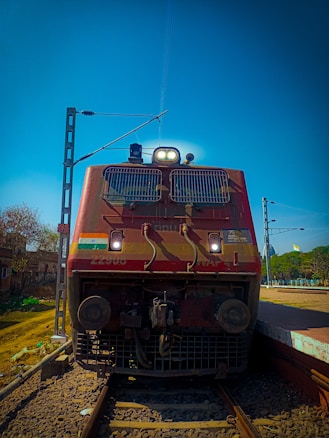A red locomotive is standing on a set of railway tracks with overhead electrical lines. It has lights on at the front and an Indian flag on the side. The scene is set at a train station with a clear blue sky and green trees in the background.