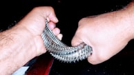 Hands gripping a shiny, metallic coil with a background of darkness. The coil consists of interlocking metal pieces, reflecting light and creating a textured appearance. The focus is on the strength and tension in the hands as they stretch and manipulate the coil.