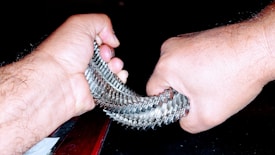 Hands gripping a shiny, metallic coil with a background of darkness. The coil consists of interlocking metal pieces, reflecting light and creating a textured appearance. The focus is on the strength and tension in the hands as they stretch and manipulate the coil.