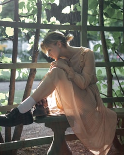 A person in a serene setting sits contemplatively on a wooden bench surrounded by leafy branches. Soft sunlight filters through the foliage, illuminating the person's face and creating a warm, tranquil atmosphere.