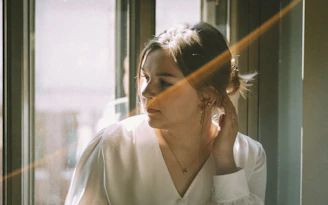 A serene woman with glowing skin sitting by a sunlit window, surrounded by soft neutral tones.