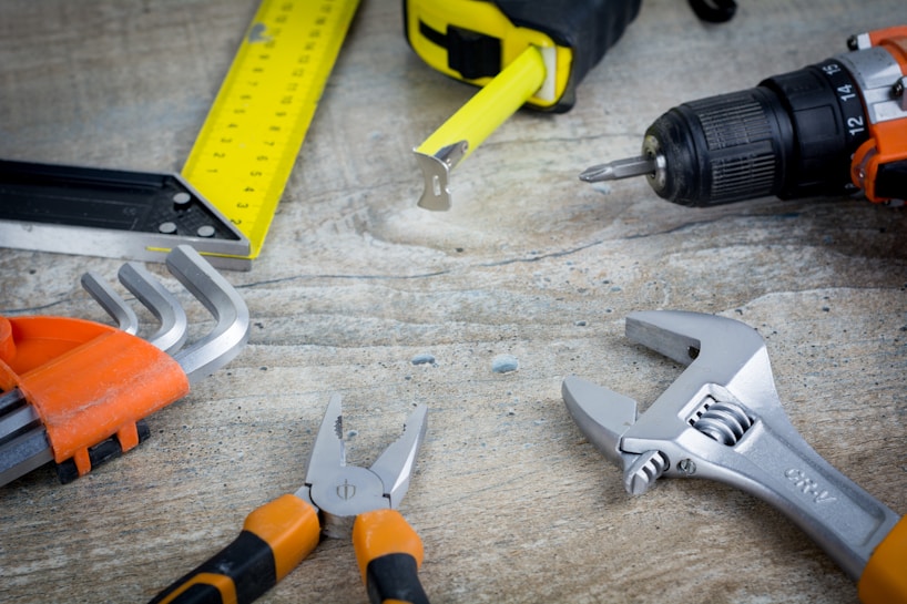 A variety of tools are laid out on a textured surface, including a yellow tape measure, a black and yellow utility knife, an orange-handled cordless drill, silver wrenches, and pliers. The items are arranged in a somewhat scattered fashion, suggesting a workspace environment.