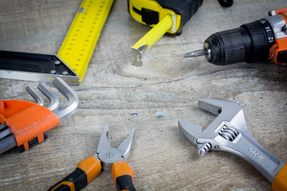A variety of tools are laid out on a textured surface, including a yellow tape measure, a black and yellow utility knife, an orange-handled cordless drill, silver wrenches, and pliers. The items are arranged in a somewhat scattered fashion, suggesting a workspace environment.