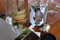 A bottle of JP. Chenet wine is placed next to a camouflage-patterned perfume bottle with a silver chain and label. Two white flowers lie nearby on a wooden surface, alongside a camera lens cap. Transparent packaging is visible in the background.