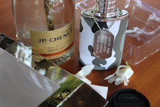 A bottle of JP. Chenet wine is placed next to a camouflage-patterned perfume bottle with a silver chain and label. Two white flowers lie nearby on a wooden surface, alongside a camera lens cap. Transparent packaging is visible in the background.