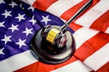 A wooden gavel with a brass band rests on a dark sound block, set against the backdrop of a United States flag. The flag features the familiar red and white stripes and white stars on a blue field, creating a strong visual association with American concepts of law and justice.