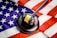 A wooden gavel with a brass band rests on a dark sound block, set against the backdrop of a United States flag. The flag features the familiar red and white stripes and white stars on a blue field, creating a strong visual association with American concepts of law and justice.