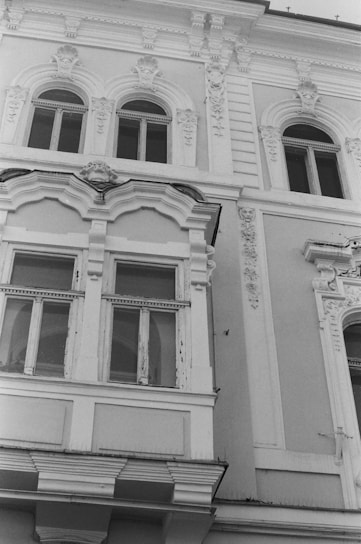 A detailed facade of a historic building featuring ornate architectural elements. The structure includes arched windows with decorative molding and intricate carvings. The windows show signs of wear, adding a touch of antiquity.