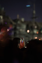 A person is holding a smartphone, capturing a photo against a dark, blurred background with bright, bokeh lights. The scene appears to be taken during a night event.
