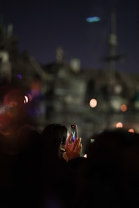 A person is holding a smartphone, capturing a photo against a dark, blurred background with bright, bokeh lights. The scene appears to be taken during a night event.