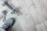 A pair of athletic shoes is next to a silver dumbbell on a light wooden floor. A water bottle with a carrying strap lies nearby.