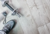 A pair of athletic shoes is next to a silver dumbbell on a light wooden floor. A water bottle with a carrying strap lies nearby.