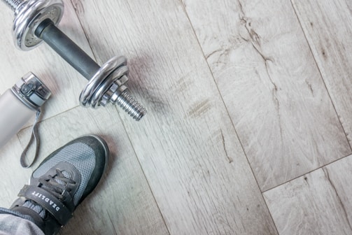 A pair of athletic shoes is next to a silver dumbbell on a light wooden floor. A water bottle with a carrying strap lies nearby.