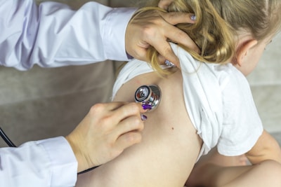 A healthcare professional uses a stethoscope to examine a child. The child has blonde hair and is wearing a white shirt, which is lifted to allow for the examination. The hands of the healthcare worker are visible, showing polished nails.