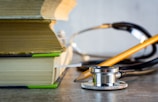 A close-up view of two stacked books with a stethoscope and a pencil resting nearby on a table. The stethoscope's metallic and rubber elements are clearly visible, and the books are thick with visible pages.