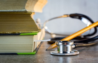 A close-up view of two stacked books with a stethoscope and a pencil resting nearby on a table. The stethoscope's metallic and rubber elements are clearly visible, and the books are thick with visible pages.