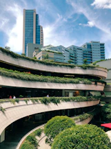 A modern urban complex with multiple levels of open terraces adorned with greenery and plants. The architecture features curving lines that create a dynamic aesthetic, while a tall glass skyscraper looms in the background against a bright blue sky.