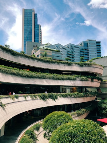 A modern urban complex with multiple levels of open terraces adorned with greenery and plants. The architecture features curving lines that create a dynamic aesthetic, while a tall glass skyscraper looms in the background against a bright blue sky.