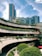 A modern urban complex with multiple levels of open terraces adorned with greenery and plants. The architecture features curving lines that create a dynamic aesthetic, while a tall glass skyscraper looms in the background against a bright blue sky.