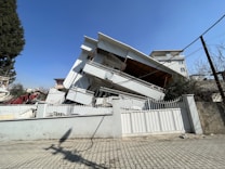 A multi-story building has collapsed, with its upper floors tilted at an angle and the structure showing significant damage. The building appears to be heavily damaged, possibly due to an earthquake or similar disaster. Surrounding buildings and infrastructure, such as the road and fences, also indicate damage.
