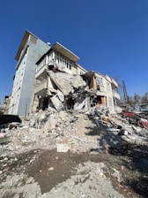 A partially collapsed multi-story residential building with significant structural damage. Debris and rubble are scattered around the base, indicative of recent destruction or disaster. The building is still standing but severely compromised, with parts of it leaning dangerously and sections of walls missing.