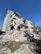 A partially collapsed multi-story residential building with significant structural damage. Debris and rubble are scattered around the base, indicative of recent destruction or disaster. The building is still standing but severely compromised, with parts of it leaning dangerously and sections of walls missing.