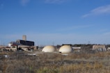 The image depicts a rural industrial setting with two large dome-shaped structures prominently in the foreground. These structures are surrounded by a complex of buildings including a tall grain elevator or silo. The area is bordered by dry grass and open land, under a clear blue sky.