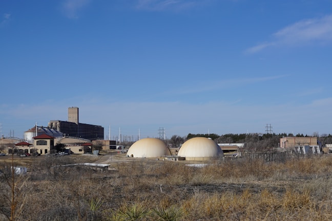 The image depicts a rural industrial setting with two large dome-shaped structures prominently in the foreground. These structures are surrounded by a complex of buildings including a tall grain elevator or silo. The area is bordered by dry grass and open land, under a clear blue sky.