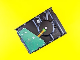 A partially disassembled hard drive is placed against a bright yellow background. The central circular component and internal green circuit board are visible.