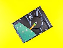 A partially disassembled hard drive is placed against a bright yellow background. The central circular component and internal green circuit board are visible.
