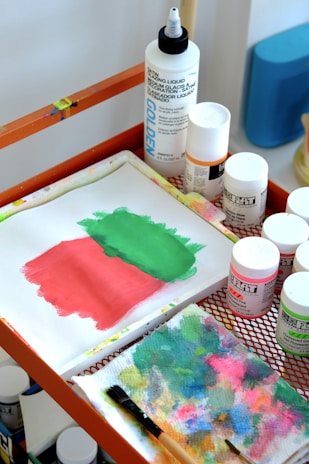 A tabletop art setup featuring a small canvas with green and pink paint, bottles of paint and medium liquid, and a colorful blotting paper with a paintbrush. The materials are organized on an orange mesh table.