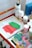A tabletop art setup featuring a small canvas with green and pink paint, bottles of paint and medium liquid, and a colorful blotting paper with a paintbrush. The materials are organized on an orange mesh table.