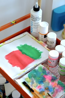 A tabletop art setup featuring a small canvas with green and pink paint, bottles of paint and medium liquid, and a colorful blotting paper with a paintbrush. The materials are organized on an orange mesh table.