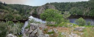 A lush, green forested area is seen surrounding a tranquil body of water, which appears to be a river or lake. The scene includes rocky outcrops and patches of grass, giving a rugged and natural appearance. Trees of various sizes fill the landscape, creating a dense canopy.