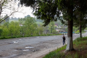 A rural area featuring a dirt road with visible potholes, bordered by lush green trees. In the background are fields and several houses, suggesting a small village or countryside setting. A person wearing a dark hoodie and jeans stands near a tree on the edge of the road.