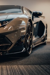 A sleek, modern sports car with a glossy finish and angular design is parked indoors. The focus is on the front end, highlighting the detailed headlight, aerodynamic curves, and stylish wheels. The lighting creates reflections and shadows, enhancing the car's luxurious appearance.