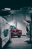 A red vintage car is parked inside a garage with subdued lighting. The car features a decorative stripe on the hood and is labeled with a badge that reads 'Supercar Club'. Nearby, other vehicles and garage equipment are partially visible.