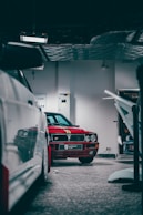 A red vintage car is parked inside a garage with subdued lighting. The car features a decorative stripe on the hood and is labeled with a badge that reads 'Supercar Club'. Nearby, other vehicles and garage equipment are partially visible.