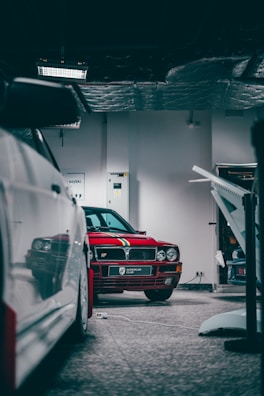 A red vintage car is parked inside a garage with subdued lighting. The car features a decorative stripe on the hood and is labeled with a badge that reads 'Supercar Club'. Nearby, other vehicles and garage equipment are partially visible.