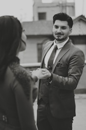 A man in a suit gently holding the hand of another person, possibly engaging in a moment of connection or conversation. The setting appears to be urban, with blurred buildings in the background. The image is in black and white, adding a classic or timeless feel.