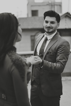 A man in a suit gently holding the hand of another person, possibly engaging in a moment of connection or conversation. The setting appears to be urban, with blurred buildings in the background. The image is in black and white, adding a classic or timeless feel.