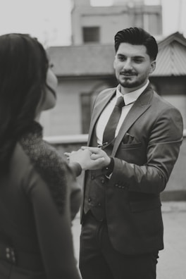 A man in a suit gently holding the hand of another person, possibly engaging in a moment of connection or conversation. The setting appears to be urban, with blurred buildings in the background. The image is in black and white, adding a classic or timeless feel.