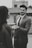 A man in a suit gently holding the hand of another person, possibly engaging in a moment of connection or conversation. The setting appears to be urban, with blurred buildings in the background. The image is in black and white, adding a classic or timeless feel.